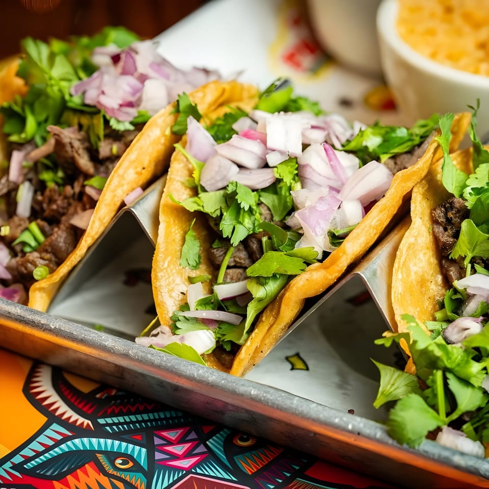 Three El Paso Mexican Grill tacos with meat, cilantro, and red onions in a metal holder.