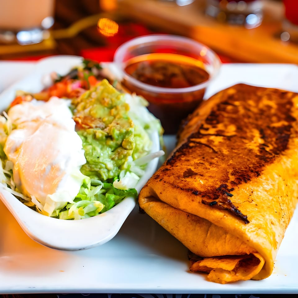A golden-brown burrito with a side of guacamole, sour cream, lettuce, and salsa on a white plate.